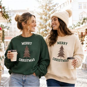 Könnte beinhalten: Ein dunkelgrünes Sweatshirt und ein cremefarbenes Sweatshirt, beide mit dem Schriftzug Merry Christmas und einer zentralen Weihnachtsbaumgrafik mit Leopardenmuster. Beide Kleidungsstücke haben lange Ärmel und einen Rundhalsausschnitt.