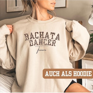 Può includere: Felpa beige con la scritta "BACHATA DANCER" in un font leopardato. Sotto la scritta, il nome "Tamara" in corsivo. Le parole "AUCH ALS HOODIE" sono in un rettangolo marrone in basso nell'immagine.
