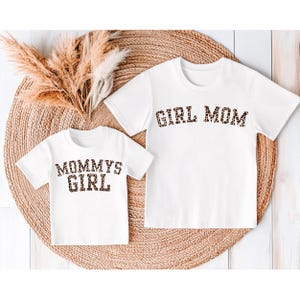 May include: Two white t-shirts with matching leopard print text. One shirt says "GIRL MOM" and the other says "MOMMYS GIRL". The shirts are displayed on a woven, circular mat with dried decorative plants.