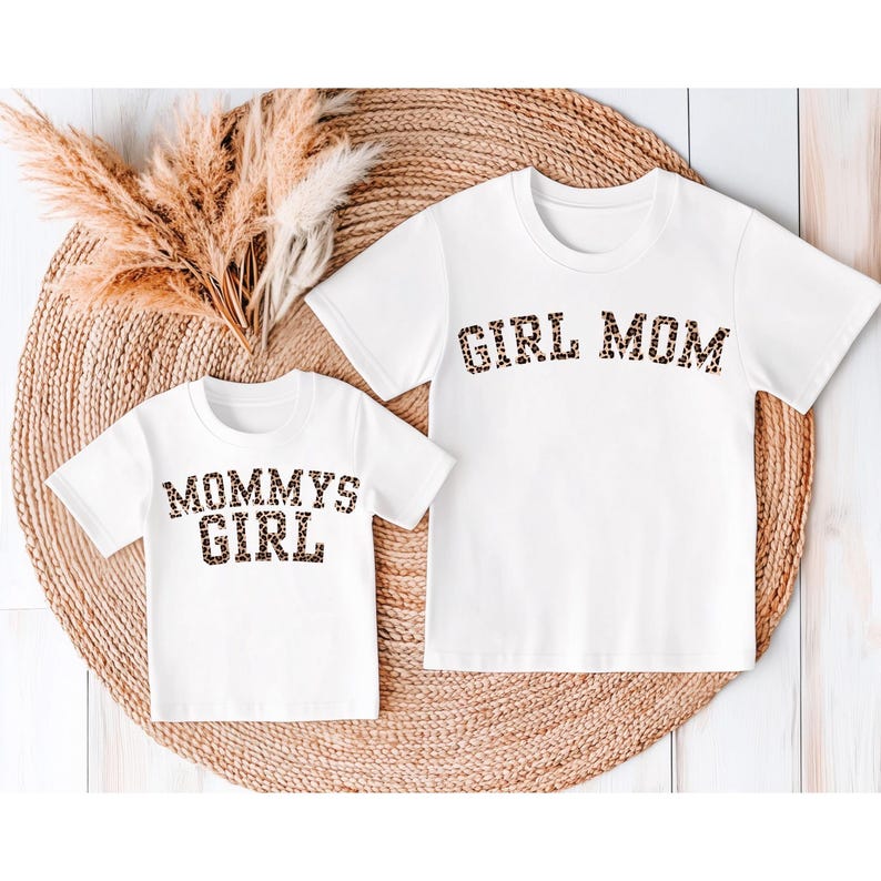 May include: Two white t-shirts with matching leopard print text. One shirt says "GIRL MOM" and the other says "MOMMYS GIRL". The shirts are displayed on a woven, circular mat with dried decorative plants.