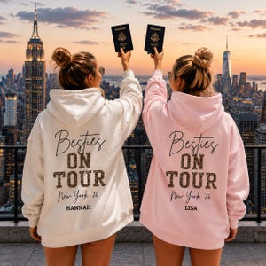 Pode incluir: Duas camisolas com capuz, uma creme e outra rosa claro, com o texto Besties ON TOUR New York '26 em preto e estampado de leopardo. A camisola creme tem o nome HANNAH impresso abaixo, enquanto a rosa tem o nome LISA impresso abaixo.