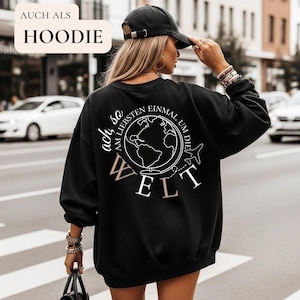 Puede incluir: Sudadera negra con un gráfico blanco de un globo terráqueo y las palabras "ach so am liebsten einmal um die Welt" y "WELT". La persona lleva una gorra negra y sostiene un bolso negro.