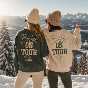 Puede incluir: Dos sudaderas de manga larga con cuello redondo, una verde oscuro y otra crema, con el texto 'Besties ON TOUR' junto a 'Ischgl 27' y un nombre impreso en la espalda. Ambas prendas se combinan con gorros de punto a juego en tonos neutros.