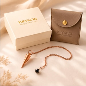 Pode incluir: Um pêndulo de metal cor de cobre com ponta afiada e uma corrente com uma pequena conta preta. O conjunto inclui uma caixa cor de creme e uma bolsa cor de taupe, ambas com o texto HRYSURI Energy Detection Tools impresso em dourado.