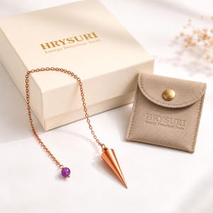 Pode incluir: Um pêndulo de metal cor de cobre com ponta afiada preso a uma corrente com uma pequena conta roxa. O conjunto inclui uma caixa creme e uma bolsa de camurça bege, ambas com o texto HRYSURI Energy Detection Tools gravado em relevo.