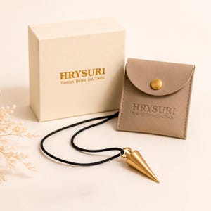 Puede incluir: Un péndulo cónico de metal dorado en un cordón negro, acompañado de una caja cuadrada beige y una bolsa de piel sintética beige a juego con un botón a presión dorado. Tanto la caja como la bolsa presentan el texto HRYSURI Energy Detection Tools.
