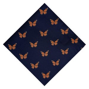 May include: Navy blue pocket square featuring a repeating pattern of orange monarch butterflies. The butterflies are detailed with intricate wing patterns, creating a striking contrast against the dark background. The fabric has a subtle ribbed texture.