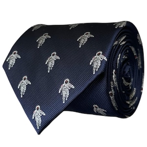 May include: A dark navy blue necktie featuring a repeating pattern of small, silver-toned embroidered astronauts. The fabric has a subtle diagonal ribbed texture, and the astronauts are depicted in a standing pose with red-tinted helmet visors.
