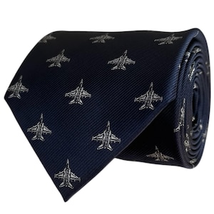 May include: A dark navy blue necktie featuring a repeating pattern of small, light grey fighter jet silhouettes. The fabric has a subtle diagonal ribbed texture, and the tie is shown rolled up to display the consistent aircraft print design.