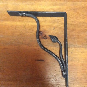 Puede incluir: Un soporte de estante de metal negro con un diseño de hoja. El soporte está hecho de hierro forjado y tiene un acabado rústico.