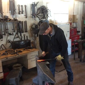 Peut inclure: Une personne portant une veste noire et un jean travaille sur une enclume en métal avec un morceau de métal rougeoyant. L'enclume est sur un support en bois. La personne utilise un marteau pour façonner le métal. Il y a des outils et du matériel accrochés au mur derrière la personne.