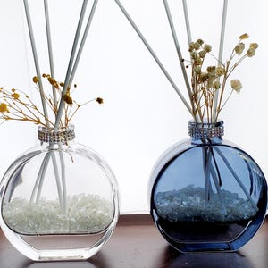 May include: Two glass aromatherapy diffusers. One is clear, the other is blue. Both have a round shape, a decorative jeweled collar, and contain white stones and reeds. Dried flowers are placed in the neck of each diffuser.