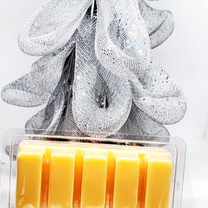 May include: A clear plastic container holding six rectangular, yellow wax melts. The melts are in front of a silver mesh decoration with glitter accents. Clear, faceted crystals are at the base of the container.