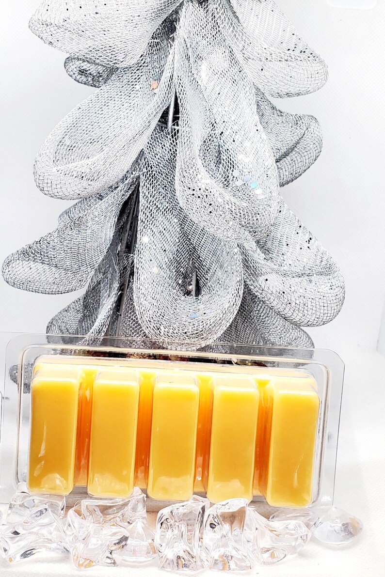 May include: A clear plastic container holding six rectangular, yellow wax melts. The melts are in front of a silver mesh decoration with glitter accents. Clear, faceted crystals are at the base of the container.