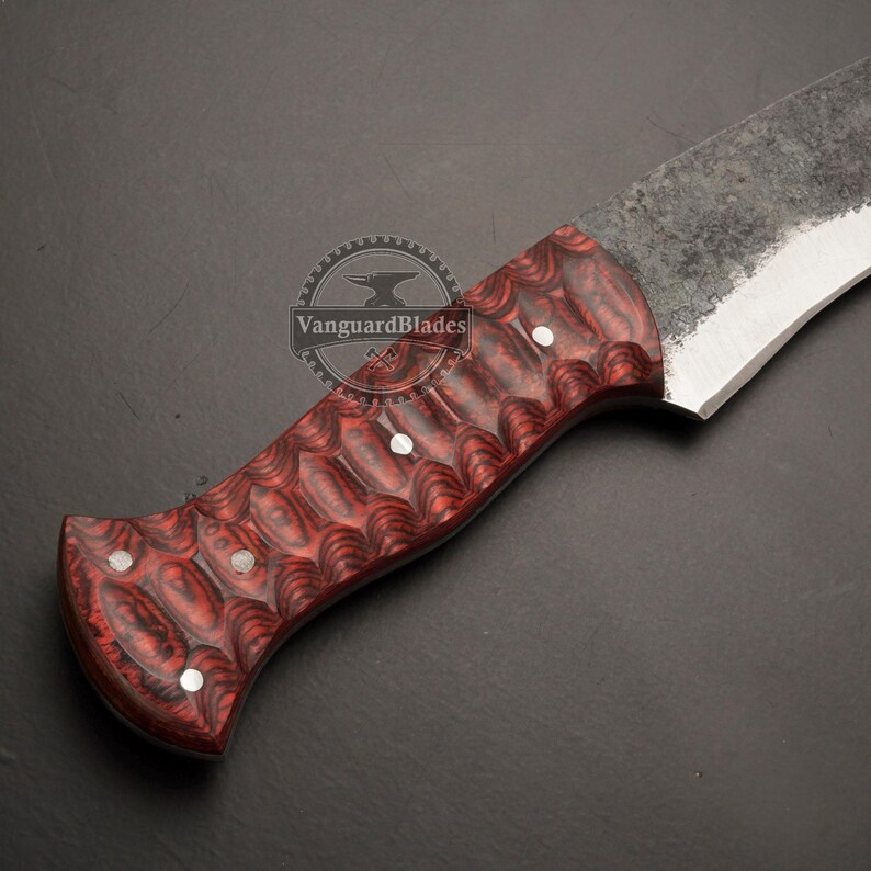 May include: Close-up of a handcrafted knife with a red and black patterned handle. The blade is a light gray color with a textured finish. The handle is secured with three silver rivets.