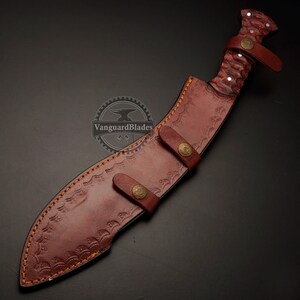 May include: A handcrafted kukri knife with a red and brown patterned handle and a brown leather sheath. The sheath features decorative stitching and two leather straps with brass-colored buttons. The knife is designed for outdoor use.