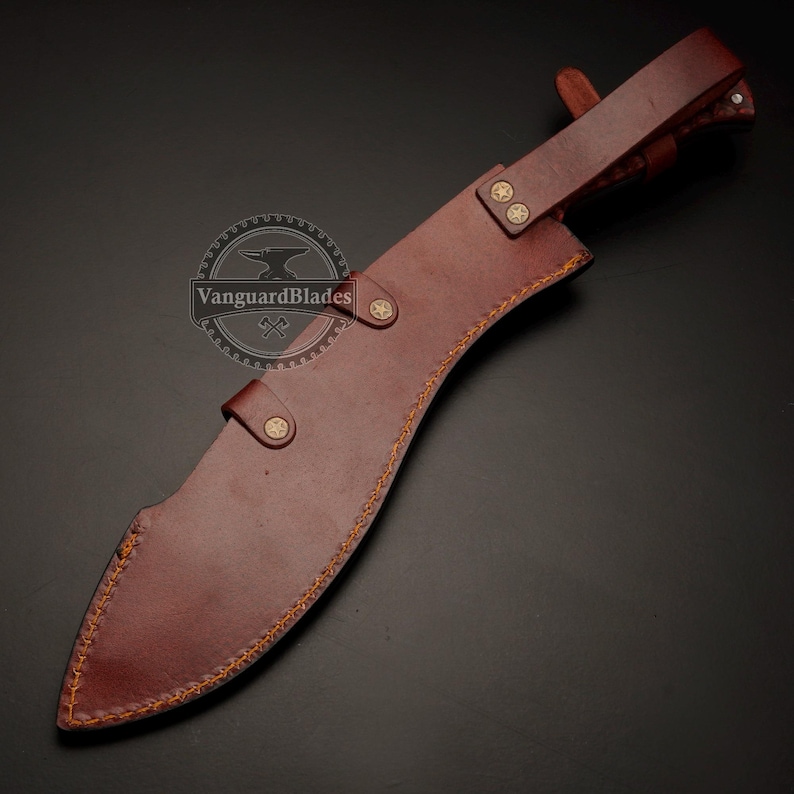May include: A brown leather sheath with a kukri knife inside. The sheath has decorative stitching and is secured with leather straps and brass-colored studs. The knife is partially visible, with a curved blade. The Vanguard Blades logo is visible.