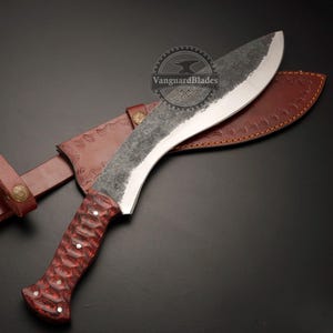 May include: A large, curved kukri knife with a textured, dark blade and a red wooden handle. The knife rests in a brown leather sheath with decorative stitching and a belt loop. The blade has a silver edge.