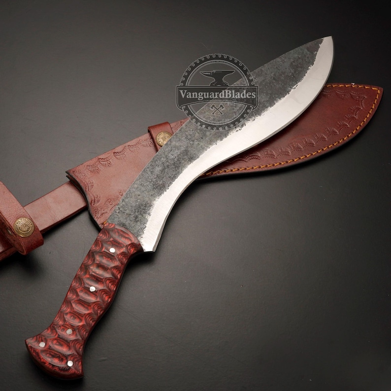 May include: A large, curved kukri knife with a textured, dark blade and a red wooden handle. The knife rests in a brown leather sheath with decorative stitching and a belt loop. The blade has a silver edge.
