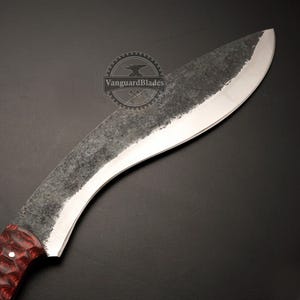 May include: Close-up of a large, curved blade knife. The blade has a textured, dark gray finish with a polished edge. The handle is a deep red with a wood grain pattern. The knife is set against a black background.