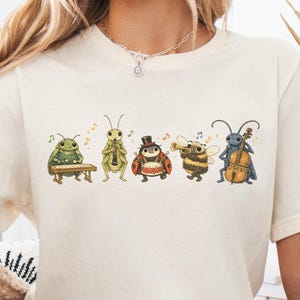 Op de afbeelding: Een natuurkleurig t-shirt met korte mouwen en een grafische print van vijf insecten die muziekinstrumenten bespelen, waaronder een kever op een xylofoon, een sprinkhaan met een klarinet, een lieveheersbeestje op een trommel, een bij met een trompet en een krekel met een cello.