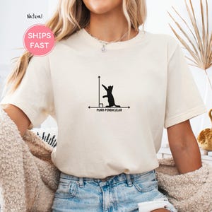 Puede incluir: Camiseta color crema con un gráfico negro de un gato en un diagrama matemático, con el texto "PURR-PENDICULAR". La camiseta tiene cuello redondo y mangas cortas. La imagen también incluye un círculo rosa con las palabras "SHIPS FAST".