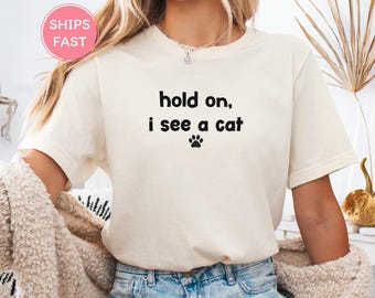 Puede incluir: Camiseta color crema con la frase negra "hold on, i see a cat" y una huella de pata. La camiseta de manga corta es de material suave. La palabra "Natural" está en la esquina superior izquierda.
