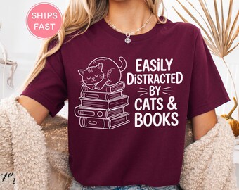 May include: A maroon t-shirt featuring a white graphic of a cat napping on a pile of books. The text on the shirt reads "Easily Distracted by Cats & Books".