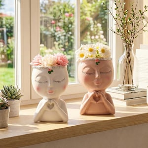 Könnte beinhalten: Zwei Keramik-Gesichtspflanzer, einer weiß und einer rosa, jeweils mit Blumen geschmückt. Die Pflanzgefäße haben geschlossene Augen und Hände in Herzform. Sukkulenten und eine kleine Tasse stehen ebenfalls auf der Fensterbank.