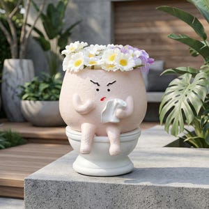 May include: A whimsical, light pink ceramic planter shaped like a person sitting on a toilet, with a grumpy face and a floral crown. The planter has arms and legs, and holds a tissue. The planter is sitting on a white toilet.