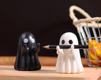 Puede incluir: Dos portalápices con forma de fantasma, uno negro y otro blanco, están sobre una tabla de madera. El fantasma blanco sostiene un lápiz. Una pequeña calabaza, una cesta de frutas y una piña están en el fondo. Accesorios de escritorio con temática de Halloween.
