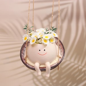 Könnte beinhalten: Ein skurriler, cremefarbener Keramik-Blumentopf in Form eines lächelnden Charakters, der in einer Schaukel sitzt. Der Topf ist mit weißen und gelben, gänseblümchenartigen Blumen und grünem Laub gefüllt. Die Schaukel ist an Schnüren aufgehängt.