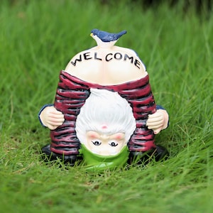 Può includere: Una statua di gnomo da giardino stravagante con barba bianca e cappello verde, piegata con la scritta "WELCOME" sulla schiena. Lo gnomo indossa un abito a righe rosse e nere e ha un uccello blu appollaiato sulla testa.
