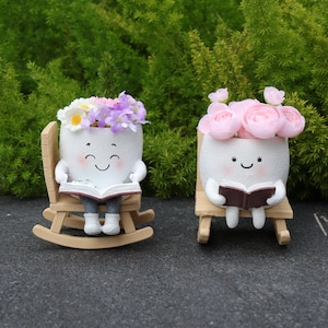 Op de afbeelding: Twee witte keramische bloempotten in de vorm van lachende personages, elk met een boek en zittend in een houten stoel. Eén pot is gevuld met kleurrijke bloemen, de andere met roze rozen. De achtergrond is groen.