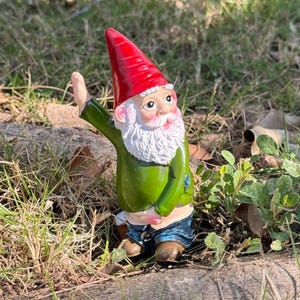 May include: A whimsical garden gnome figurine with a red hat, white beard, and green shirt. The gnome is waving with one hand and has a mischievous expression on its face.