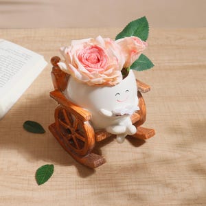 Puede incluir: Una maceta de cer&aacute;mica blanca caprichosa con forma de personaje leyendo un libro, ubicada en una mecedora de madera. La maceta contiene rosas rosas y hojas verdes. La silla de madera tiene un acabado marr&oacute;n.