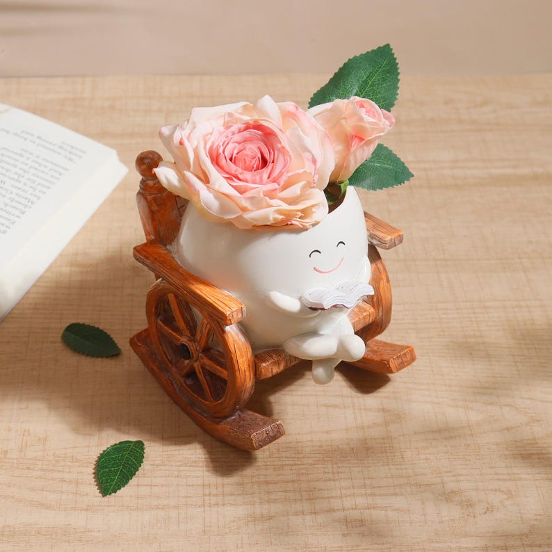 Puede incluir: Una maceta de cer&aacute;mica blanca caprichosa con forma de personaje leyendo un libro, ubicada en una mecedora de madera. La maceta contiene rosas rosas y hojas verdes. La silla de madera tiene un acabado marr&oacute;n.
