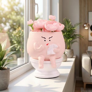 Pode incluir: Um vaso de cerâmica rosa caprichoso em forma de personagem sentado em uma privada, segurando um lenço. O vaso é preenchido com flores rosas. O personagem tem uma expressão de raiva. O vaso está em uma janela.