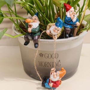 Peut inclure: Un pot de plante décoré de trois figurines de gnomes. Les gnomes portent des tenues bleues, rouges et vertes avec des barbes et des chapeaux blancs. Un gnome se balance sur une corde, et le pot porte l'inscription "GOOD MORNING".