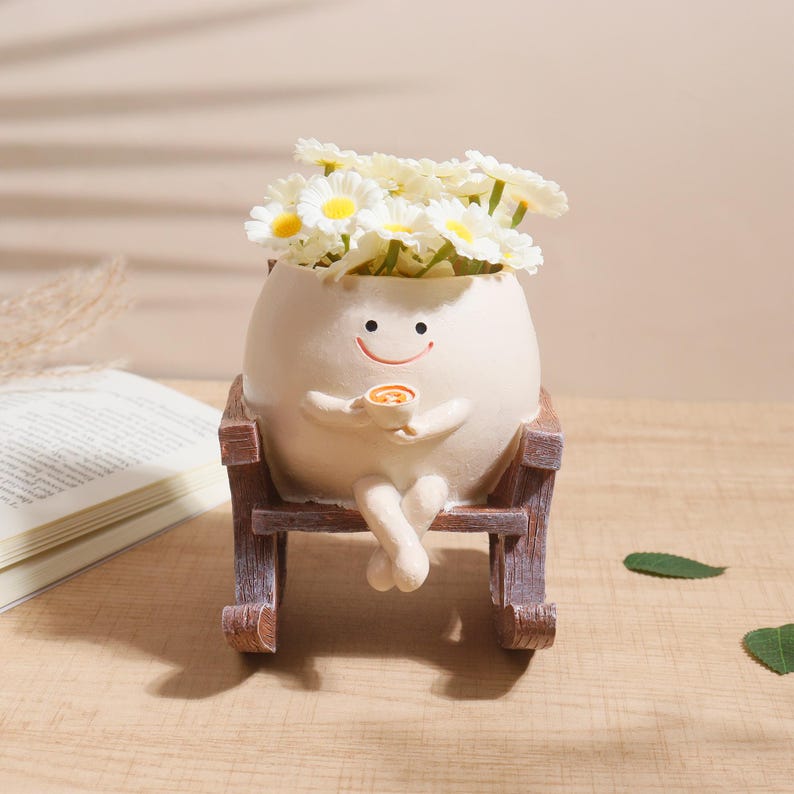 Puede incluir: Una maceta caprichosa en forma de huevo con una cara sonriente, sosteniendo una taza y sentada en una mecedora de madera. La maceta est&aacute; llena de flores blancas parecidas a margaritas con centros amarillos. Una pieza decorativa encantadora.
