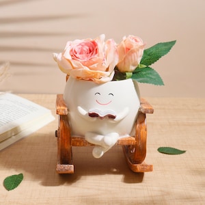 May include: A white ceramic planter in the shape of a character reading a book, set in a wooden rocking chair. It contains peach-coloured roses and green leaves. The design is charming and decorative.