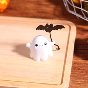 May include: A white ghost figurine with a smiling face and pink cheeks, holding a black bat decoration. The Halloween-themed decoration is on a wooden cutting board.