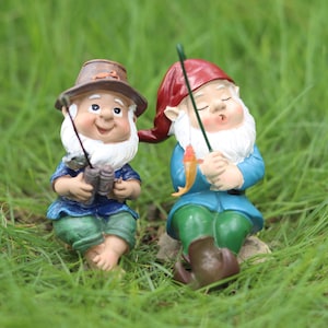 Może przedstawiać: Dwa krasnale ogrodowe, jeden w brązowym kapeluszu i niebieskiej koszuli, trzymający lornetkę i wędkę. Drugi krasnal ma czerwony kapelusz i niebieską koszulę, trzymając wędkę z małą pomarańczową rybą. Obaj mają białe brody.