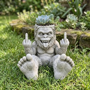 May include: A gray concrete Bigfoot planter with a succulent plant in its head. The Bigfoot is sitting with its tongue out and giving the middle finger with both hands. The planter is on a grassy lawn.