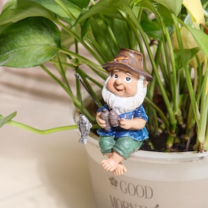 Może przedstawiać: Figurka gnoma ogrodowego, ubrana w niebieską koszulę, zielone spodnie i brązowy kapelusz, łowiąca na małą wędkę. Gnom ma białą brodę i trzyma lornetkę. Figurka siedzi na krawędzi białej doniczki.