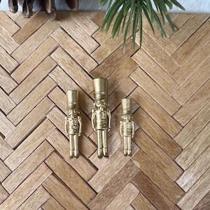 Puede incluir: Tres figuras doradas de cascanueces de diferentes tamaños están dispuestas sobre una superficie de madera. Los cascanueces están detallados con uniformes y sombreros de estilo militar. El fondo incluye una ramita de pino y una piña.