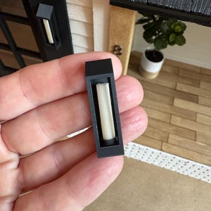Op de afbeelding: Een miniatuur zwarte buitenmuurlamp met een rechthoekige vorm, met een wit, cilindrisch licht. De lamp is ontworpen voor een poppenhuis of miniatuursc&egrave;ne, met een realistische uitstraling. Het licht is niet verlicht.