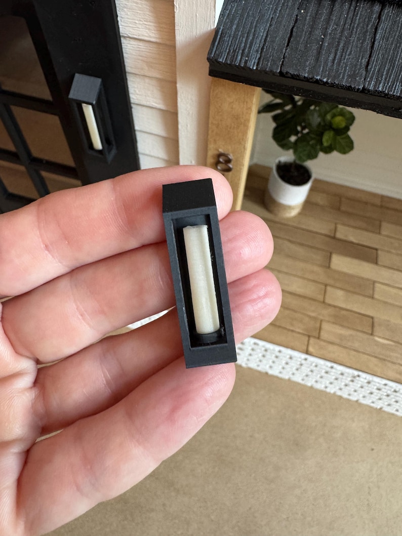 Op de afbeelding: Een miniatuur zwarte buitenmuurlamp met een rechthoekige vorm, met een wit, cilindrisch licht. De lamp is ontworpen voor een poppenhuis of miniatuursc&egrave;ne, met een realistische uitstraling. Het licht is niet verlicht.