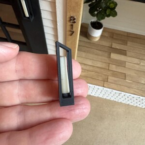 Op de afbeelding: Een kleine, rechthoekige zwarte buitenlamp met een witte binnenkant, vastgehouden in een hand. De lamp is een miniatuurmodel, waarschijnlijk voor een poppenhuis of architectuurmodel. De achtergrond bevat een miniatuurhuisgevel.