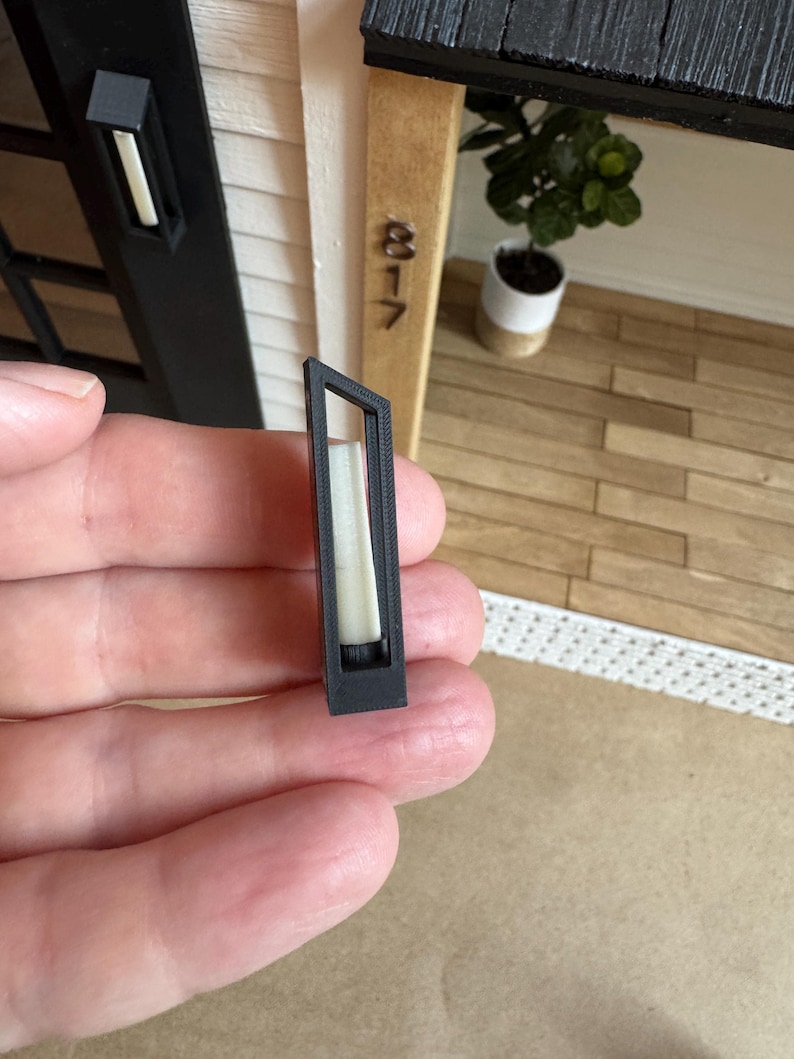 Op de afbeelding: Een kleine, rechthoekige zwarte buitenlamp met een witte binnenkant, vastgehouden in een hand. De lamp is een miniatuurmodel, waarschijnlijk voor een poppenhuis of architectuurmodel. De achtergrond bevat een miniatuurhuisgevel.